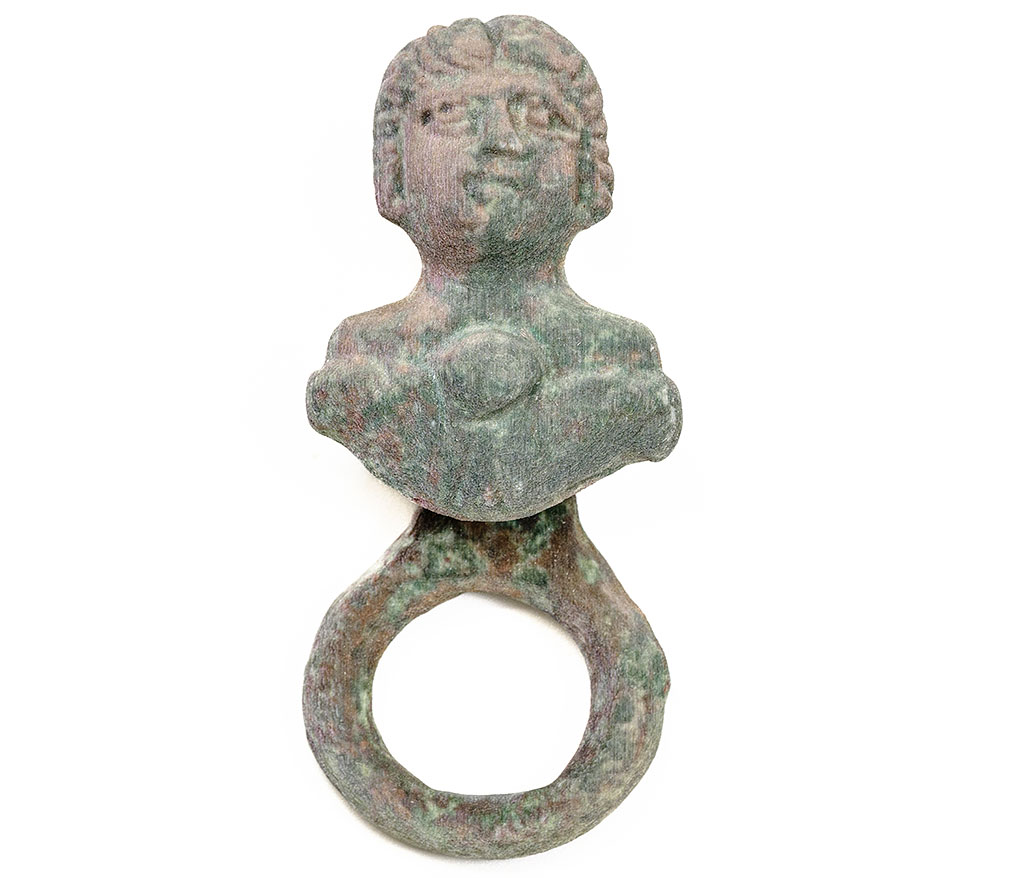 This dress fastener is cast in copper alloy and shows a goddess holding out a basket containing either a cake or a piece of fruit