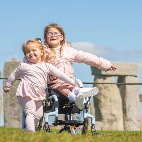 Two young girls are in front of Stonehenge, one is sitting in a wheelchair and the other stands beside her. They are both wearing pink and smiling. 
