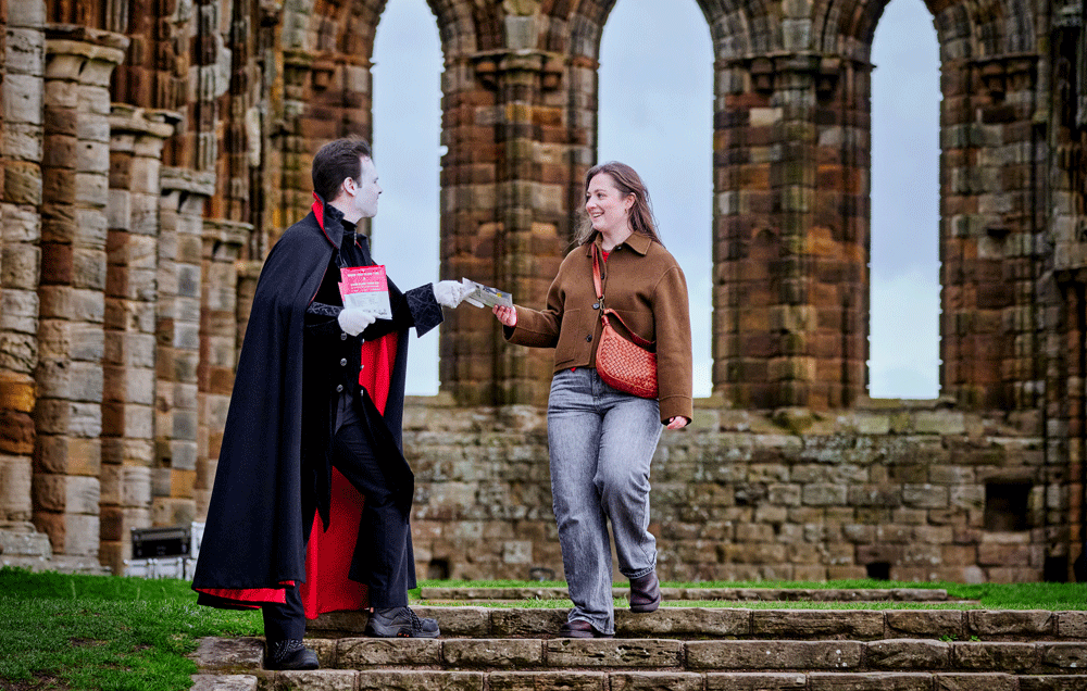 Image: an actor dressed as Dracula hand out a blood type test kit to a visitor at Whitby Abbey