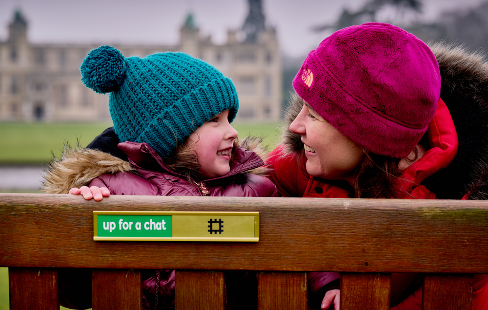 Image: a mother and daughter look at the slider on a bench which reads 'up for a chat'