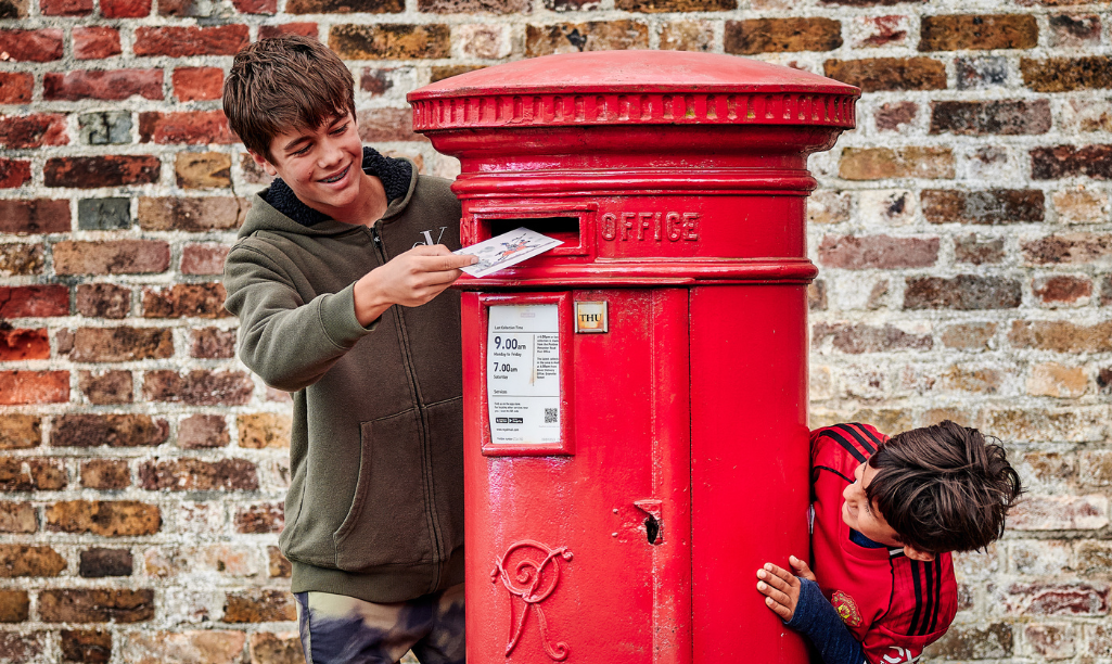 A boy posts a postcard into a post box whilst another boy looks on.