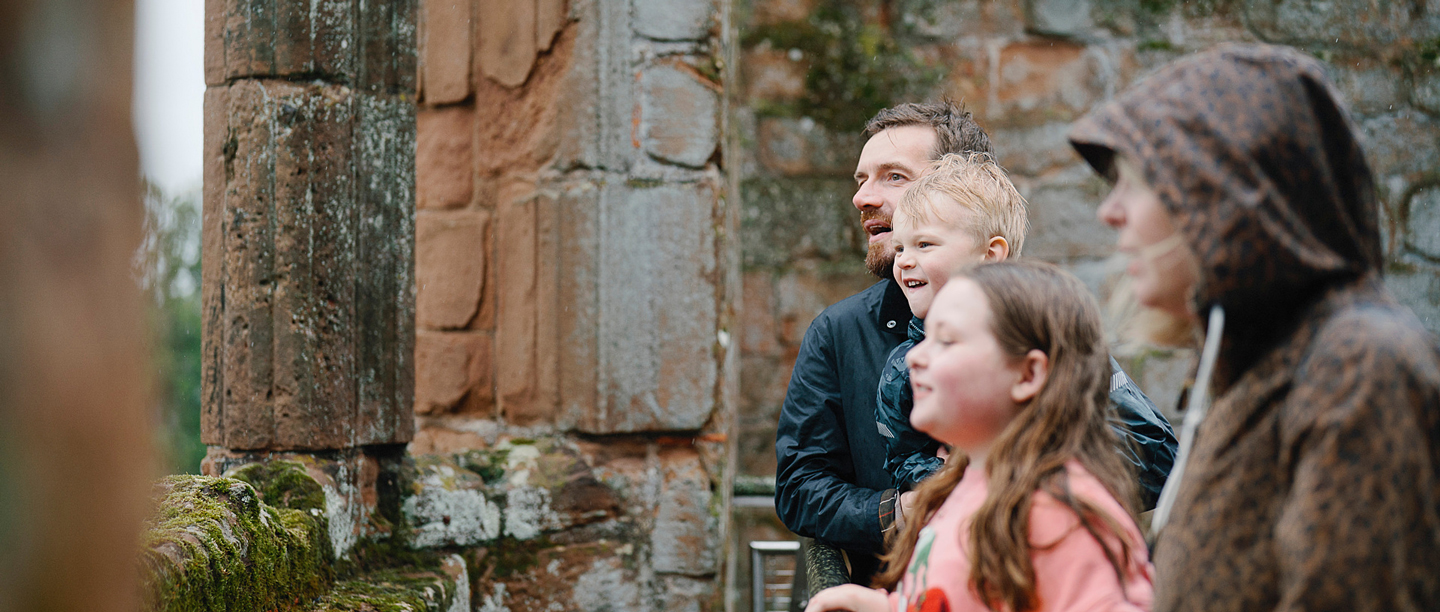 A man and a woman look out of the ruined large windows at Kenilworth Castle with two young children. They are wearing waterproof coats and jumpers.