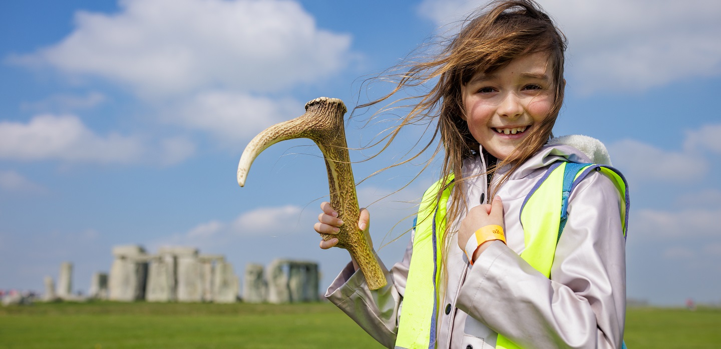 A young girl in a high vis jacket holds a replica neolithic tool in front of Stonehenge. 