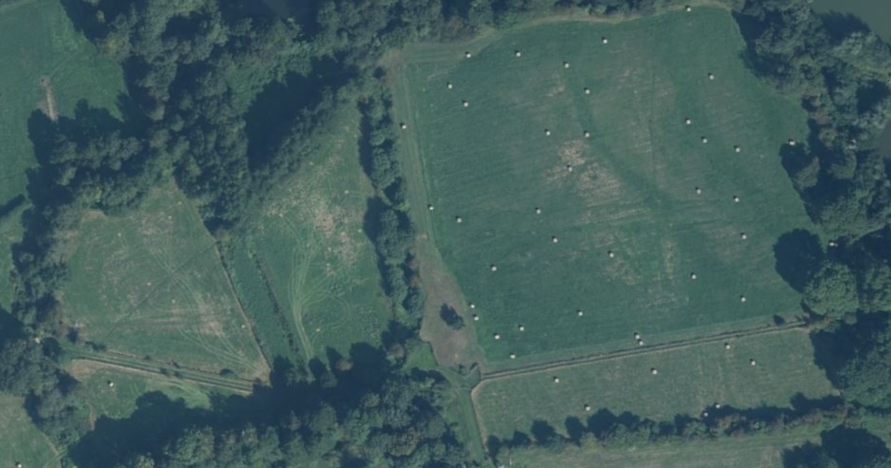 An aerial photograph showing two fields next to each other. The field on the left is surrounded by trees and has faint outlines of tracks running through it. The field on the right has a faint outline of a track running from the top of the field to the bottom.