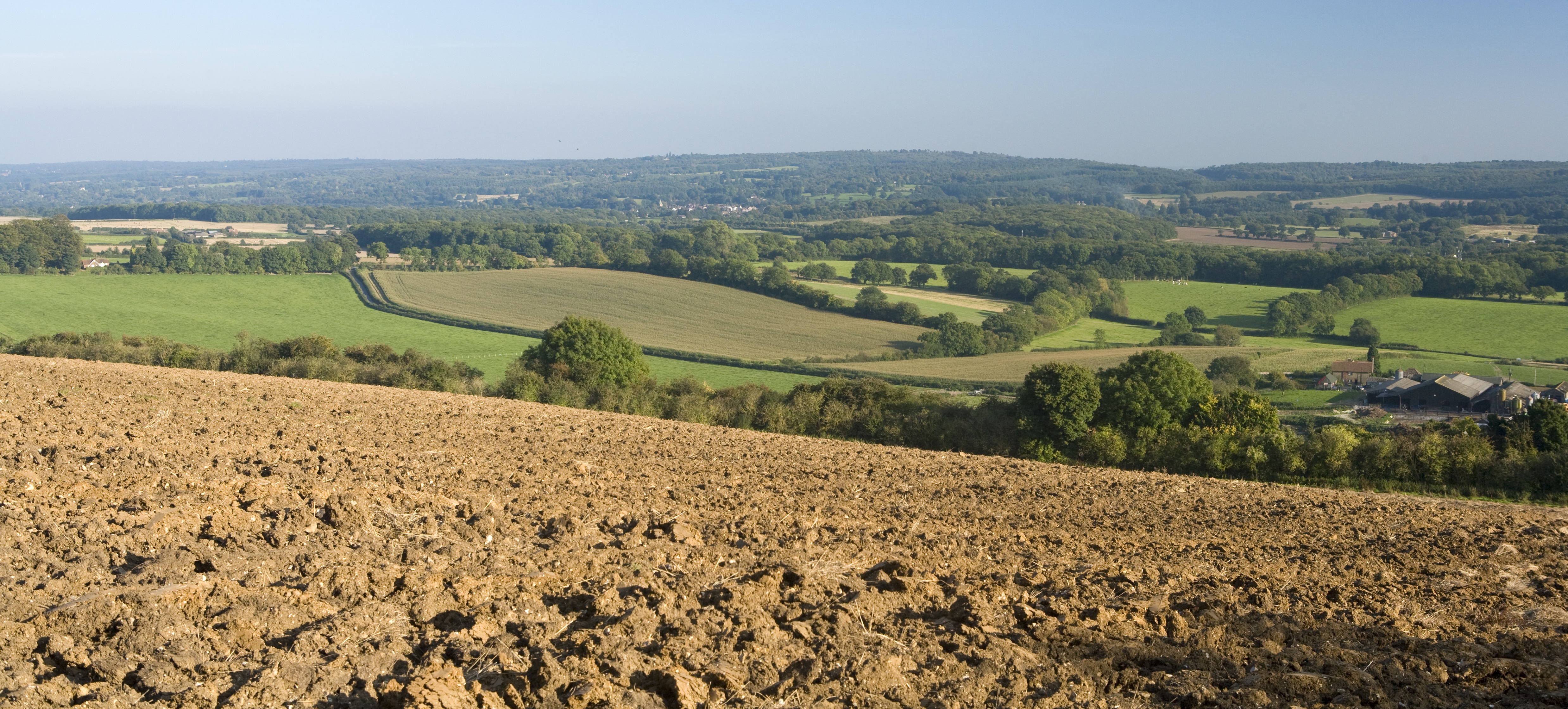 A ploughed field with the rolling hills and fields of the Darent Valley behind underneath a bright blue sky. 