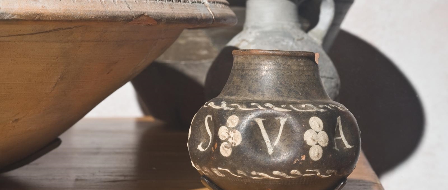 A orange deep-dish bowl next to a smaller brown glazed jug with white inscription 'SVA'. Behind this there is a grey-coloured jug with a handle on its long neck.