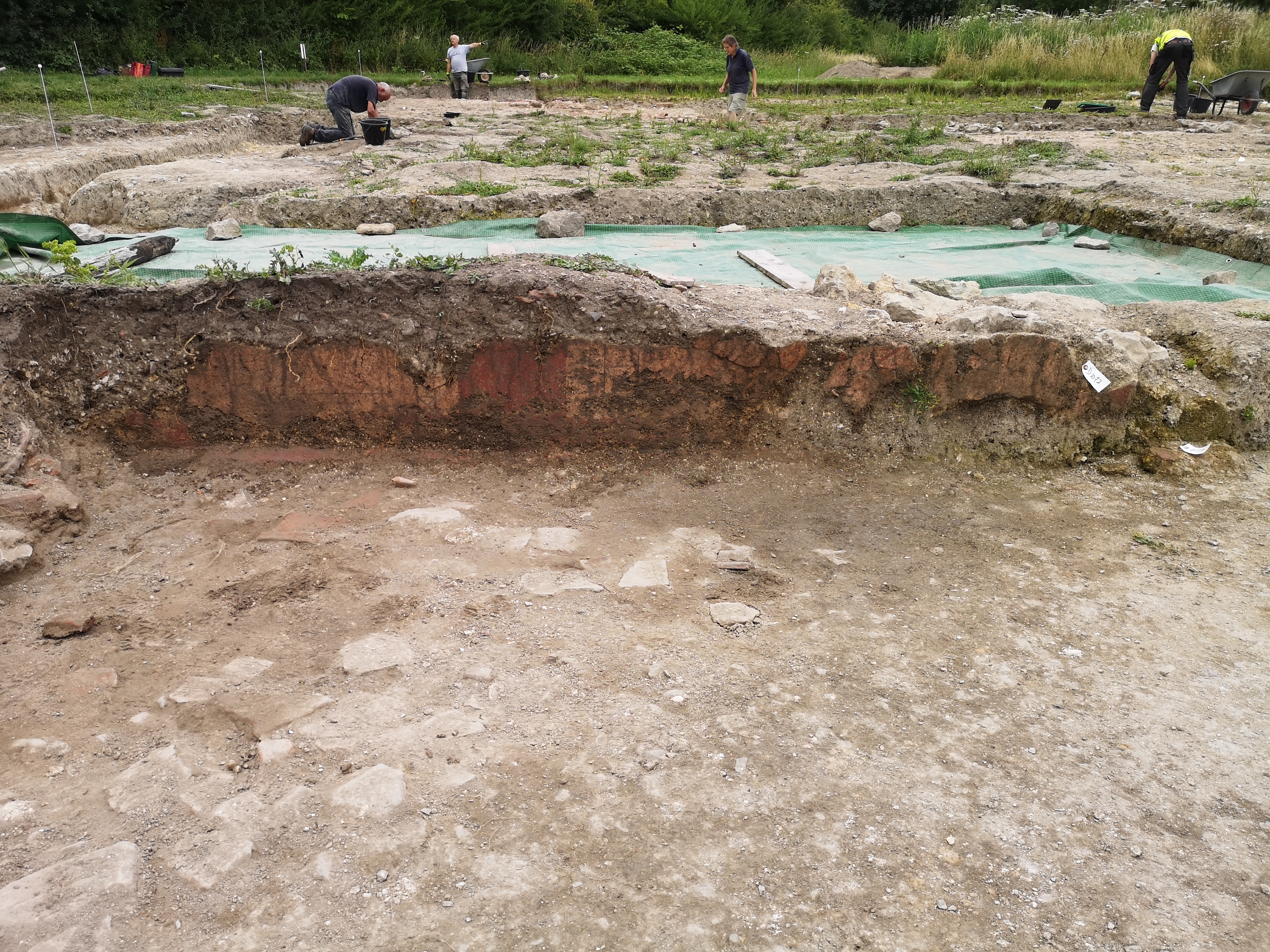 A long piece of orange and red wall plaster standing upright in an archaeologist's trench. There is an uneven stone floor in front of the plaster and excavators can be seen at ground level in another trench beyond the plaster.