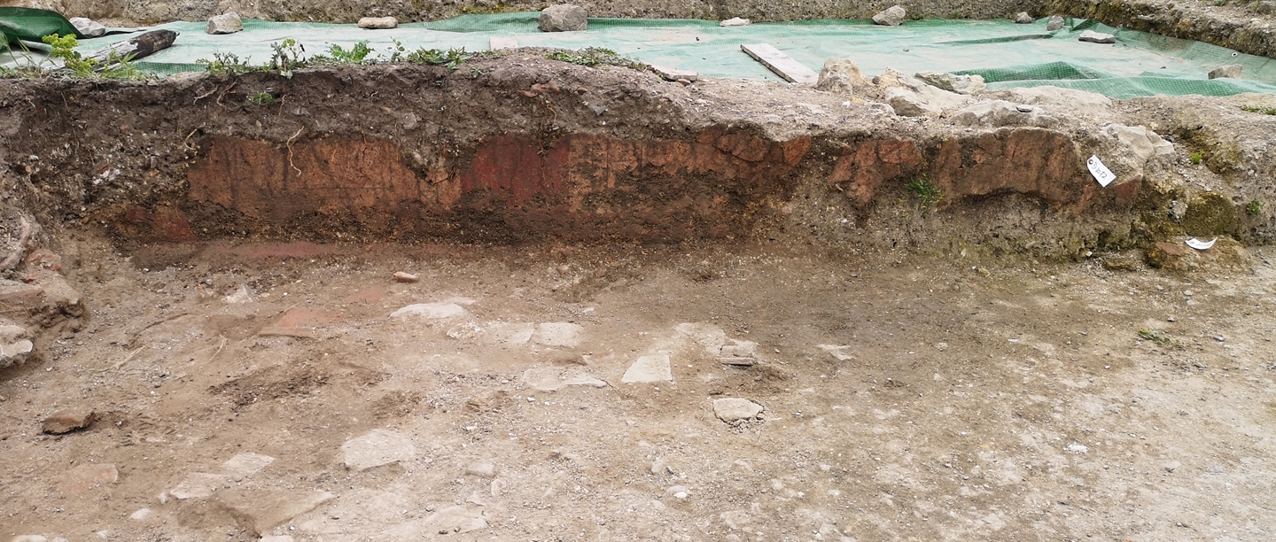 A long piece of orange and red wall plaster standing upright in an archaeologist's trench. There is an uneven stone floor in front of the plaster and excavators can be seen at ground level in another trench beyond the plaster.