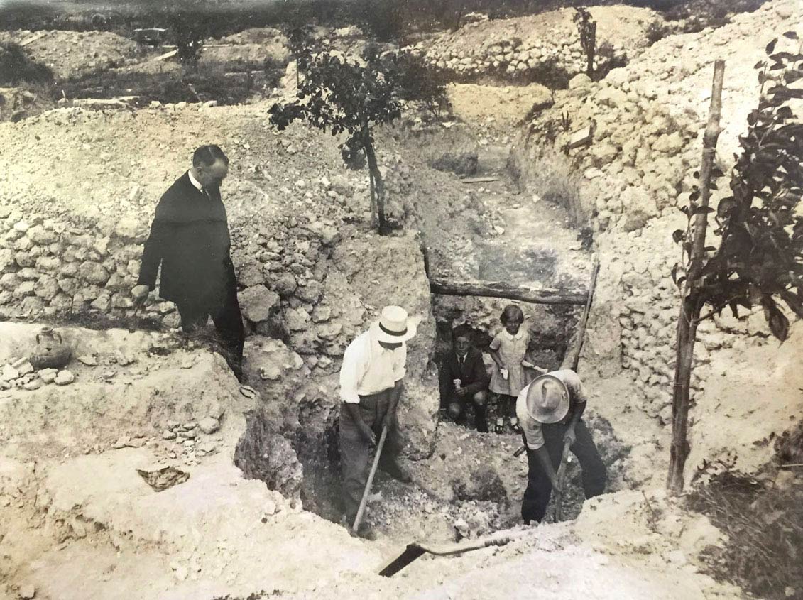 A sepia-coloured photograph of three men, a boy and a girl, inside a trench surrounded by stone villa remains.