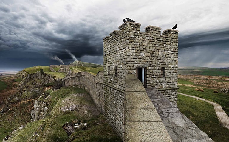 A crenellated stone tower sitting on a stone wall. The wall is located on top of a natural crag which can be seen on the left-hand side. Three dark-coloured birds sit on the top of the tower. 