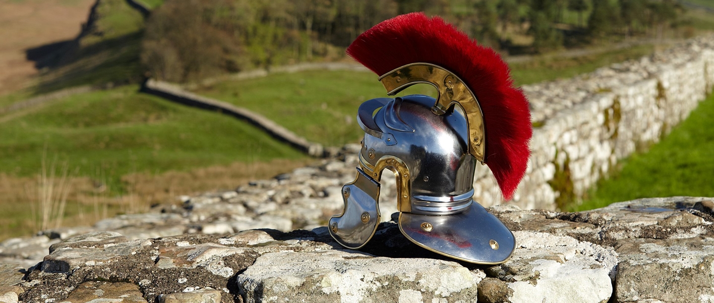 A metal Roman helmet with a dark red feather coming out of the top rests on a section of stone wall.