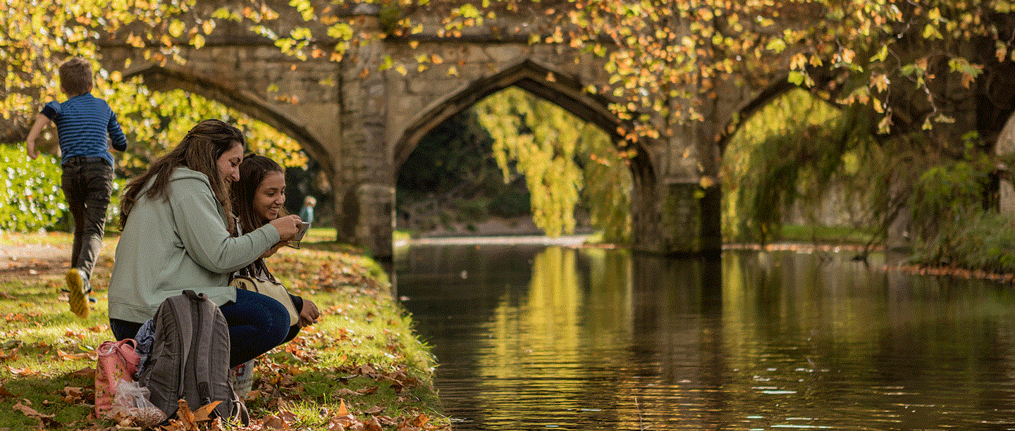 Two people kneel down by the river at Eltham Palace, smiling. There is a stone bridge in the background surrounded by autumn leaves.
