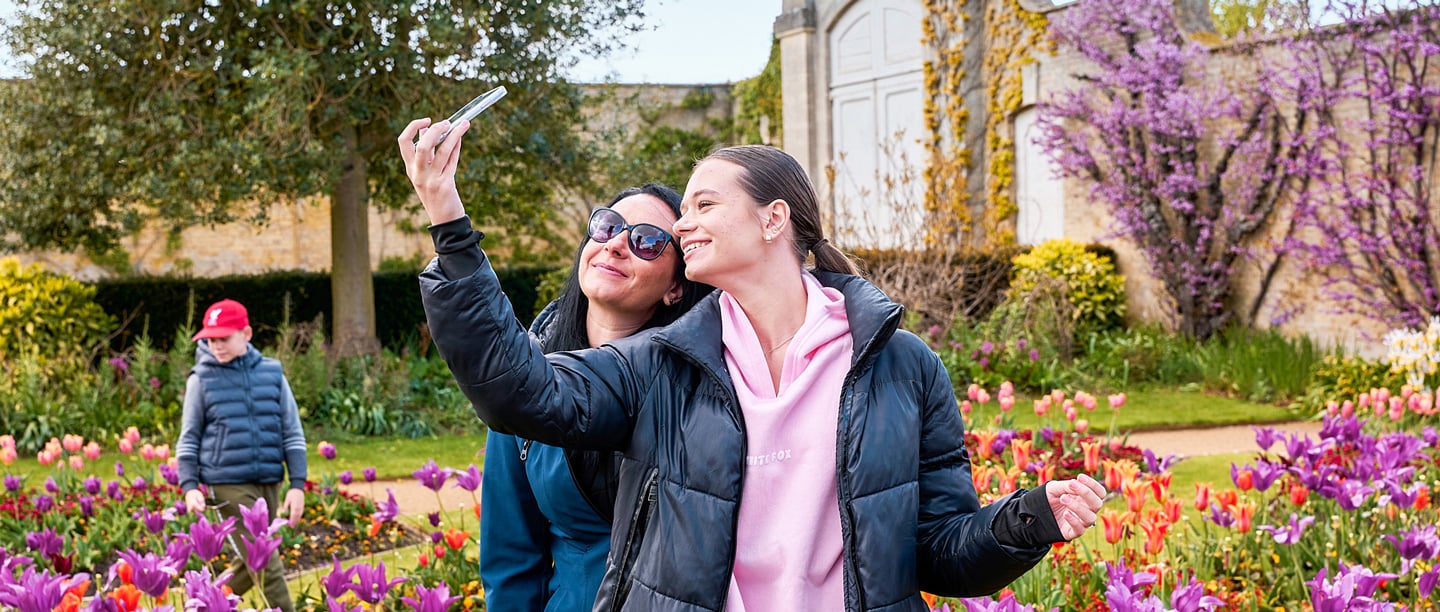 A woman and a girl take a selfie smiling, next to colourful formal gardens. A younger boy walks in the gardens behind them.