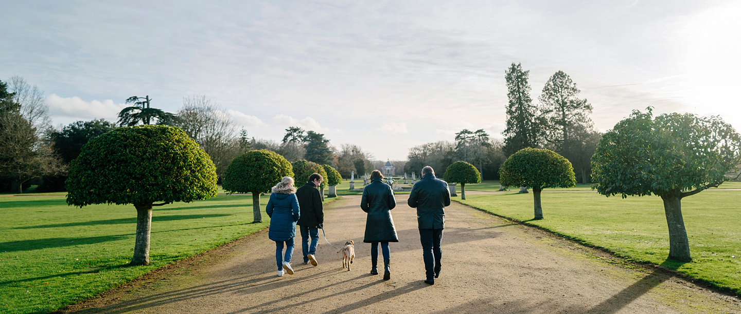 A group of four adults wearing winter coats walk a dog side by side in the gardens at Wrest Park.