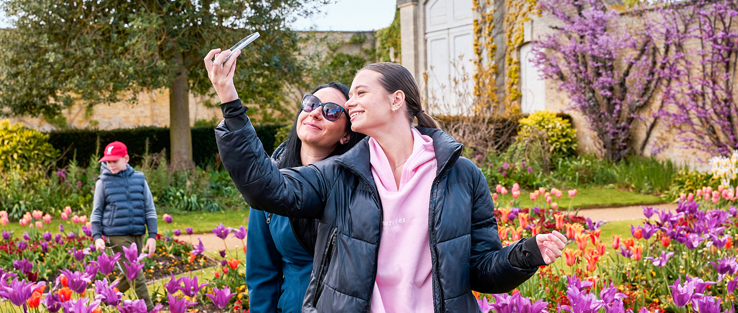 A woman and a young adult take a selfie in the gardens at Wrest Park next to the colourful flower beds.