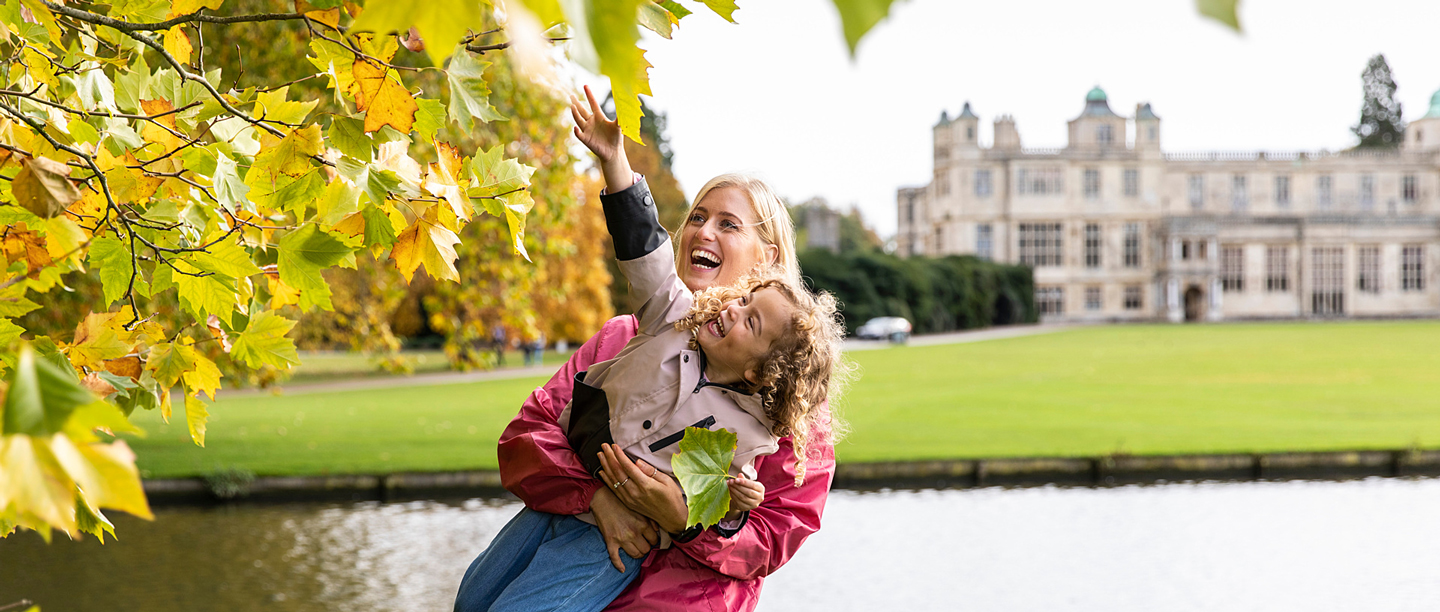 A woman lifts a child up to touch the brown leaves on a tree with Audley End House in the background.
