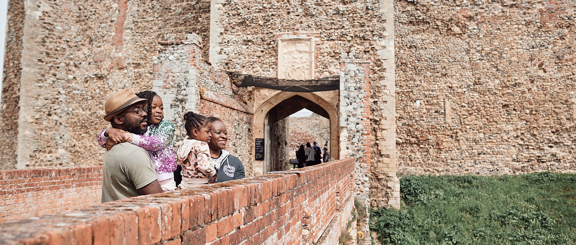 Two adults and children look over the stone bridge at Framlingham Castle on a sunny day.