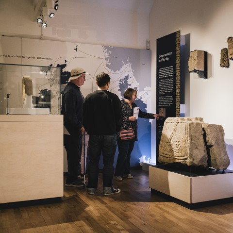 Image: Photo of three people reading an information panel in the museum at Lindisfarne Priory