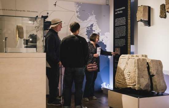 Image: Photo of three people reading an information panel in the museum at Lindisfarne Priory