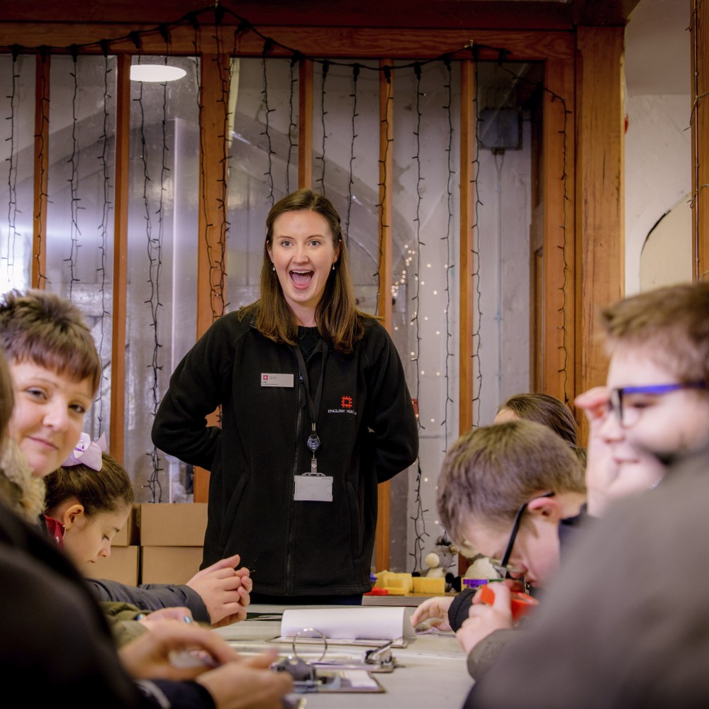 Photo of an English Heritage staff member smiling at a group of children sitting round a table at Carisbrooke Castle