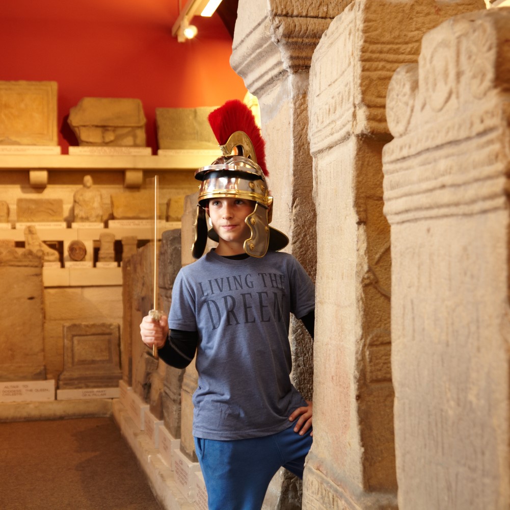 Photo of a child wearing a Roman soldier's helmet and holding a wooden sword in the museum at Chesters Roman Fort