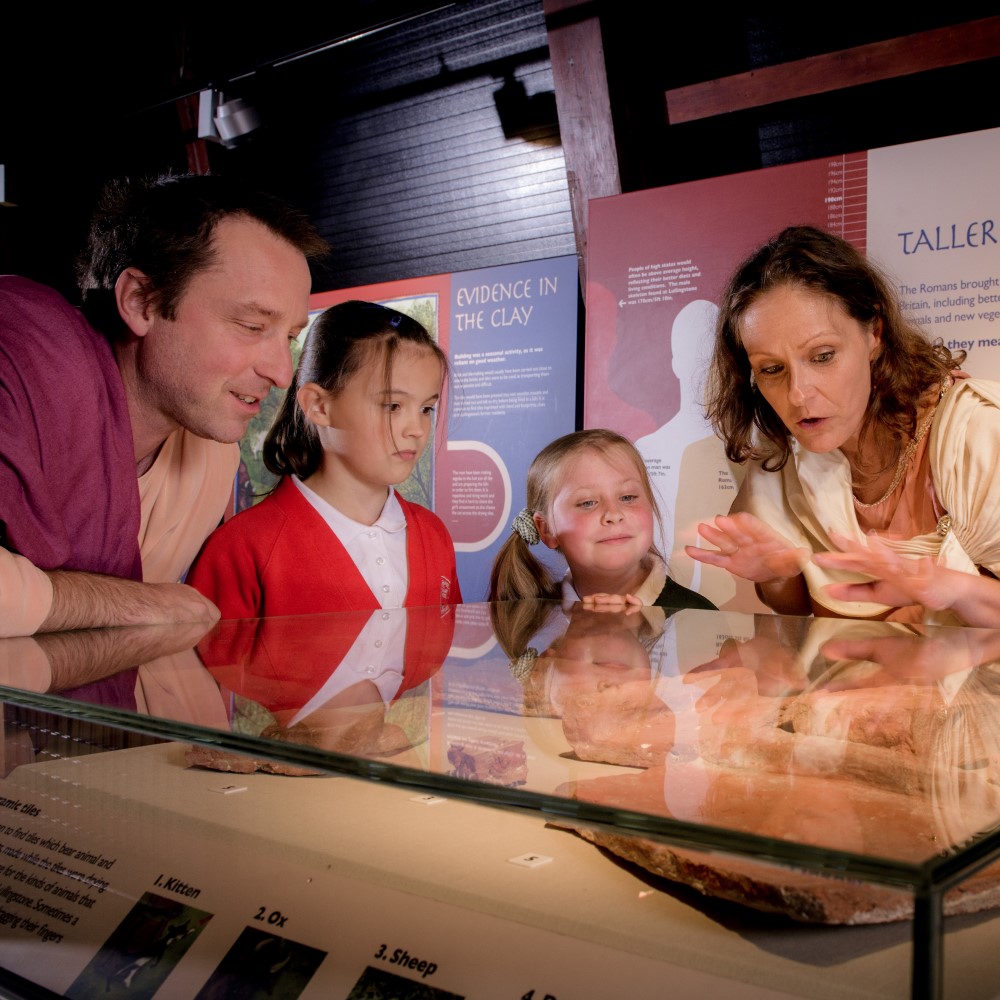 Photo of two adults in Roman costume showing two children Roman artefacts in a glass case at Lullingstone Roman Villa