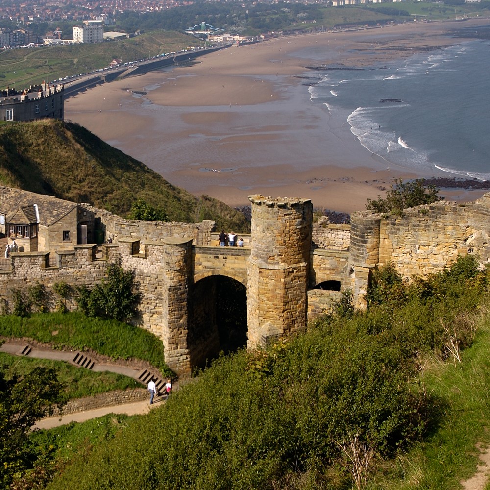 Photo of people walking around the gatehouse of Scarborough Castle with the beach and town in the background