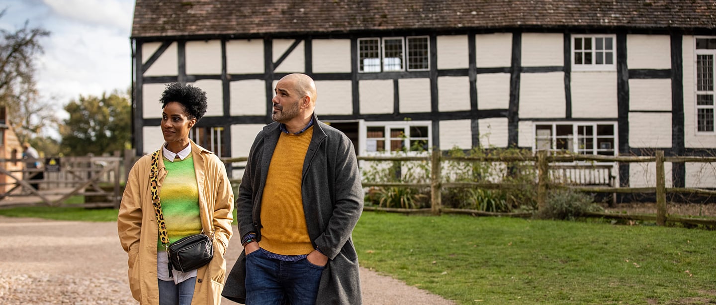Two adults walk through the gardens in front of the timber-framed Boscobel House wearing long coats and jumpers.