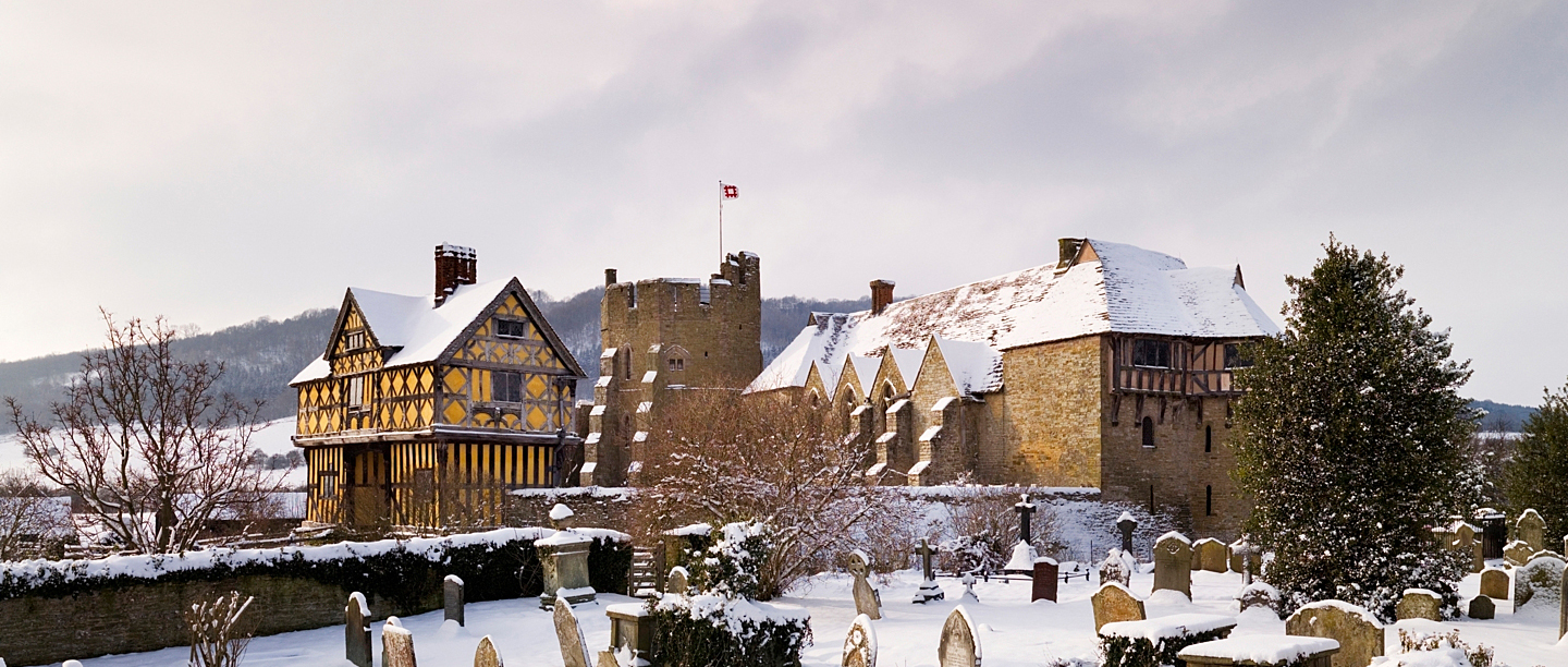 The yellow timber framed gatehouse at Stokesay Castle in the snow with the graveyard in the foreground.