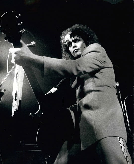 Black and white three quarter length portrait of Marc Bolan clutching a guitar with a stage light shining down on him. Bolan wears a suit with a leopard-print stripe down the side of the leg.