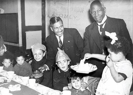 Black and white photograph of pastor Kamal Chunchie pouring tea from a pot into the cup of a child at a table, while another adult serves them a platter of sandwiches. Some of the children pull faces and wear paper hats.