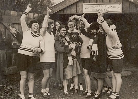 Black and white photograph of cheering women outside a boatshed