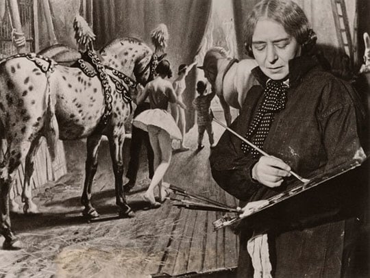 Black and white photograph of artist Laura Knight holding her paintbrushes in front of a painting of circus performers and horses