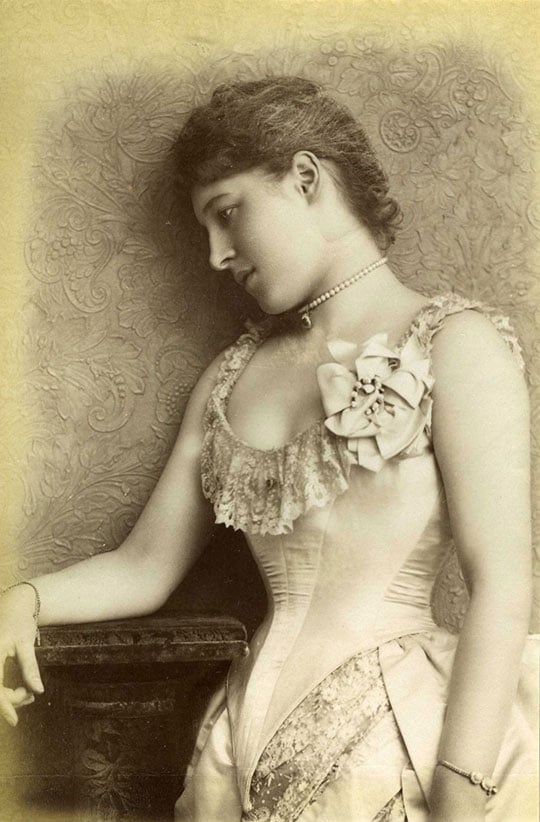 Monochrome photograph of Lillie Langtry leaning against a wall with a floral motif. She wears a pearl choker and bracelets at each wrist, and a corset top with lace trim and a fabric flower corsage