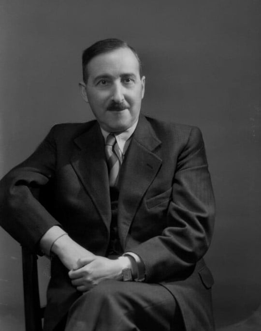 Black and white photograph of Stefan Zweig wearing a dark suit, seated with his legs crossed, his hands resting in his lap and a slight smile on his face