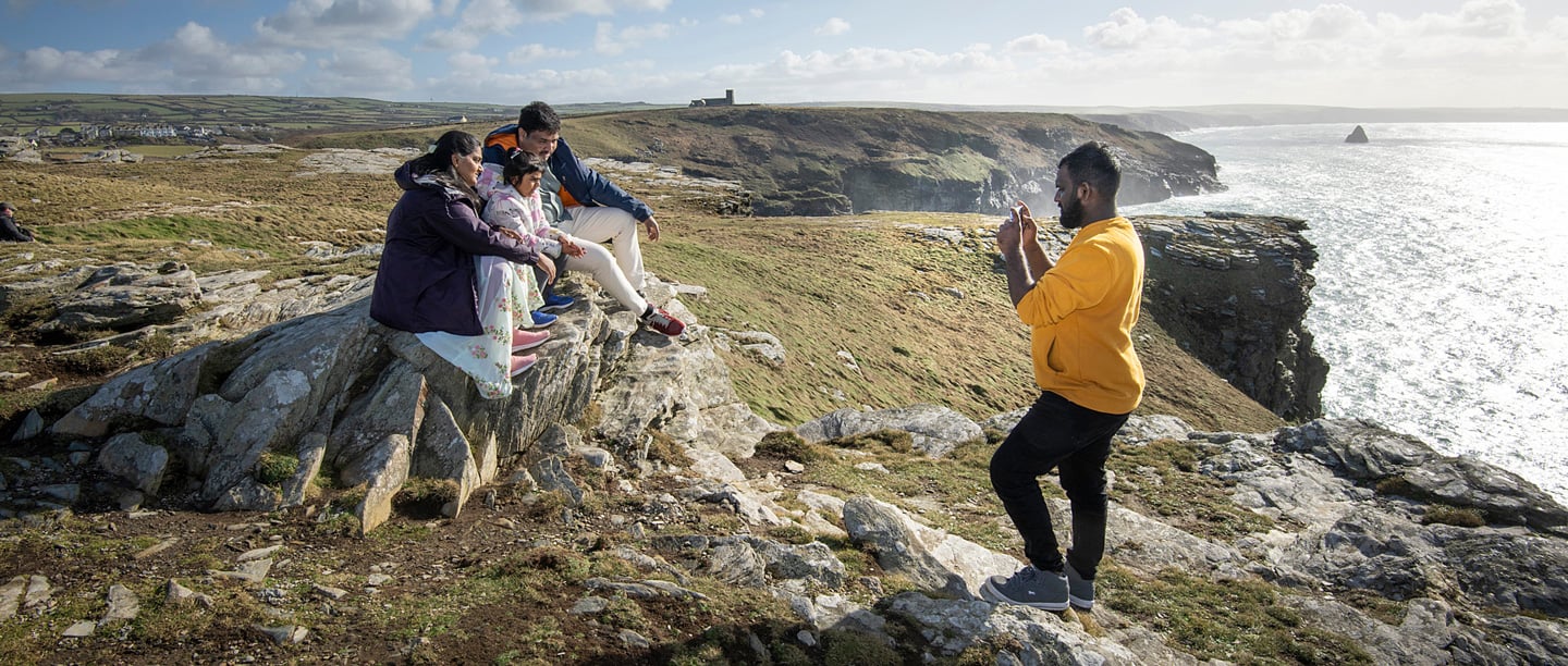 A group of adults and children sit on stones at Tintagel Castle and pose for a photo.