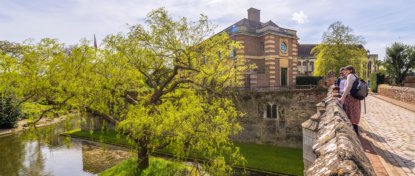 Two adults and two young children stand on the bridge at Eltham Palace overlooking the moat on a sunny day.