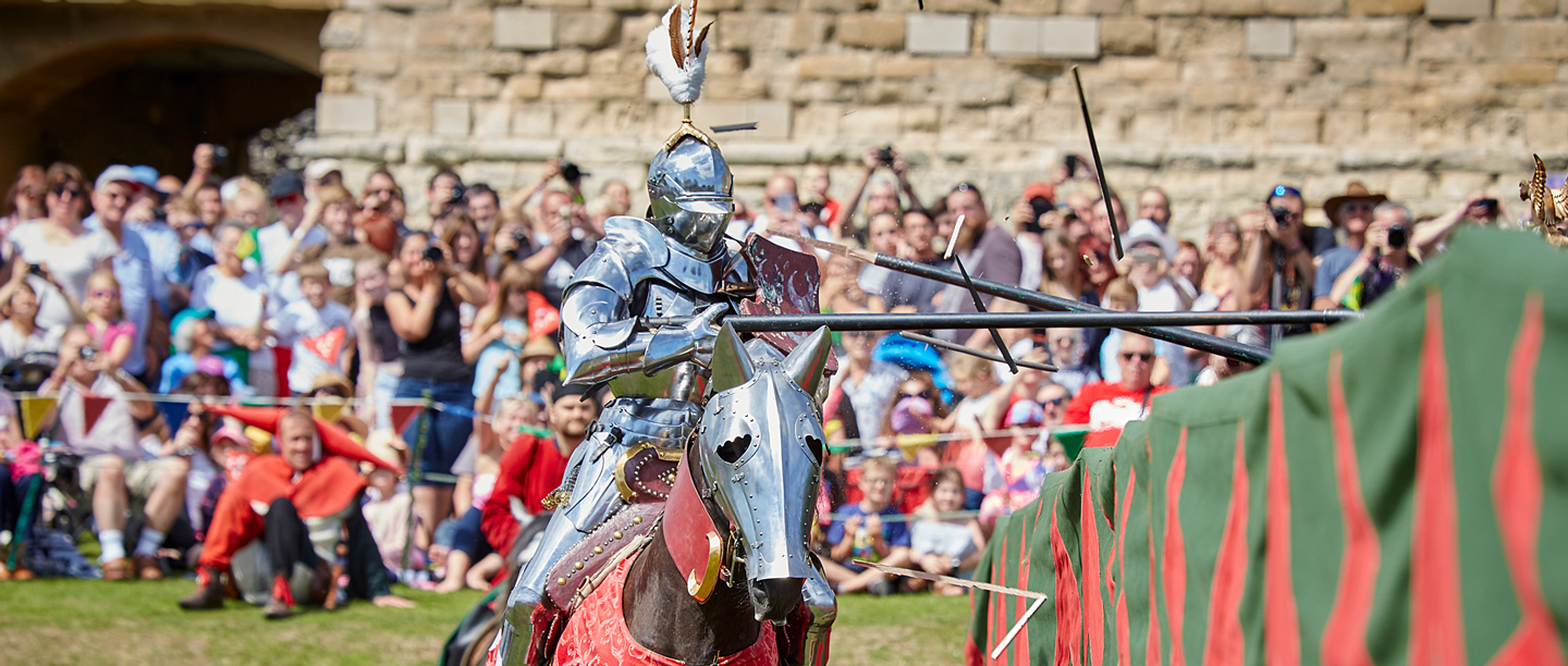 A man dressed in knights armour on horseback re-enacts a joust in front of a crowd.