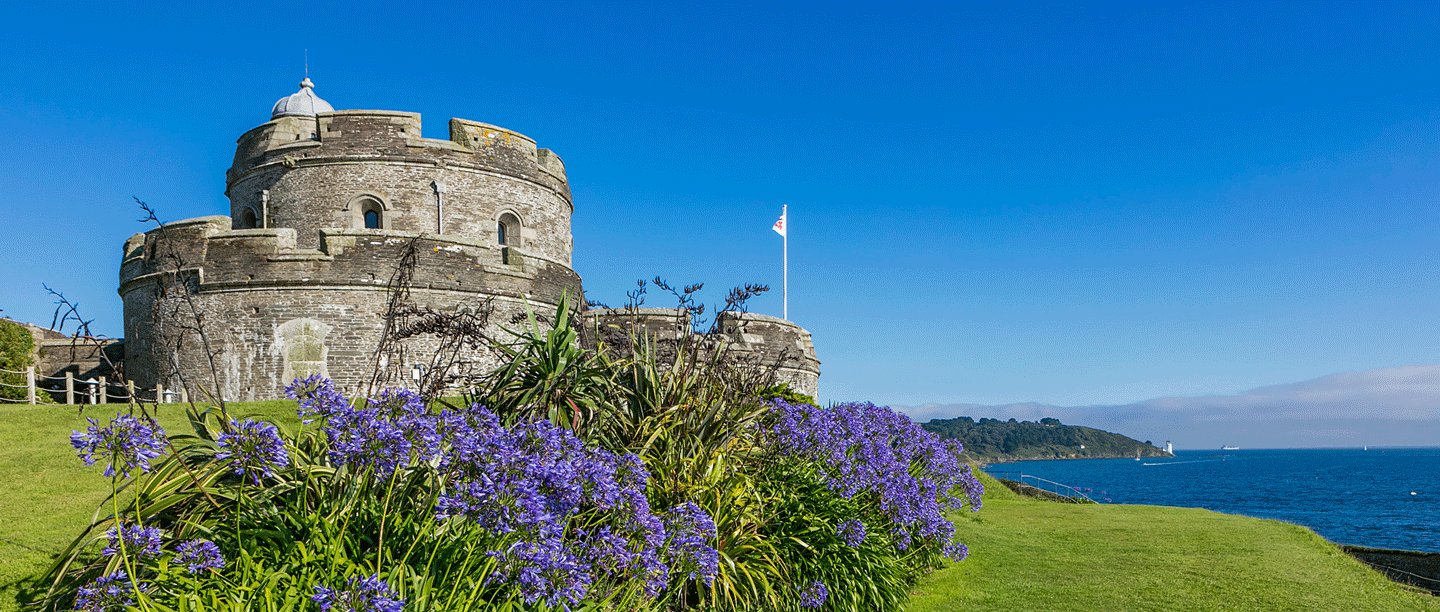 A view of an  artillery fort by the coast. The sky is blue and the castle is surrounded by purple flowers and greenery.