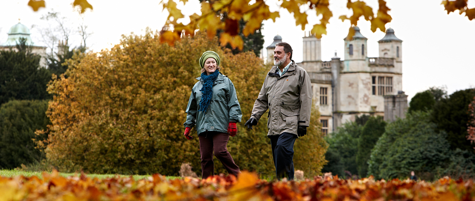 A man and a woman walk through gardens with autumn leaves on the tree and on the floor.
