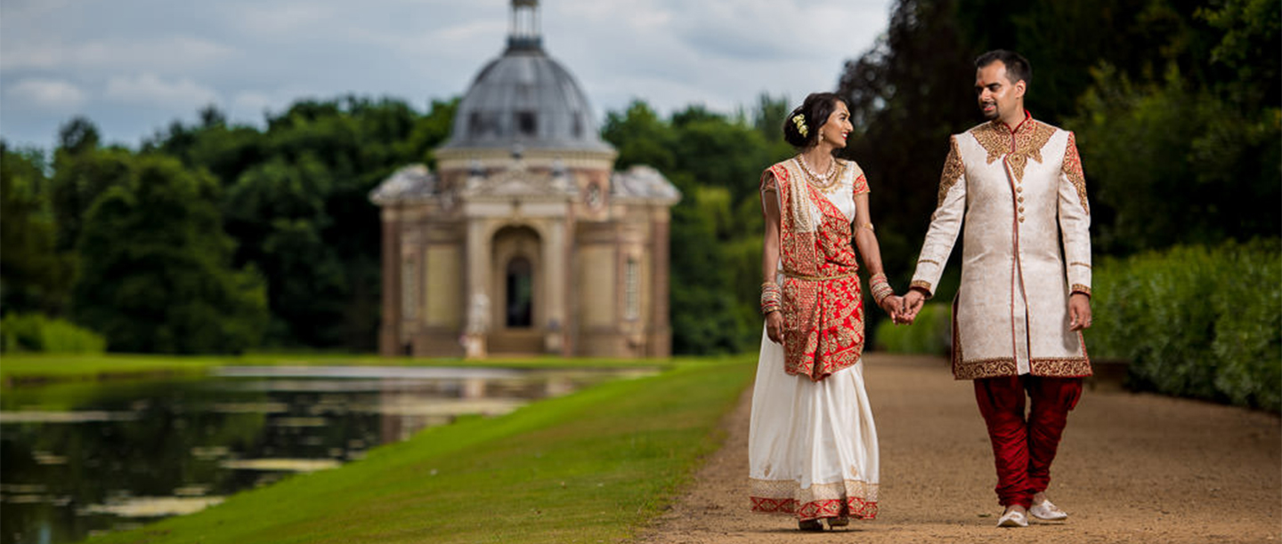 A couple in wedding finery walk down a path with a grand pavilion behind