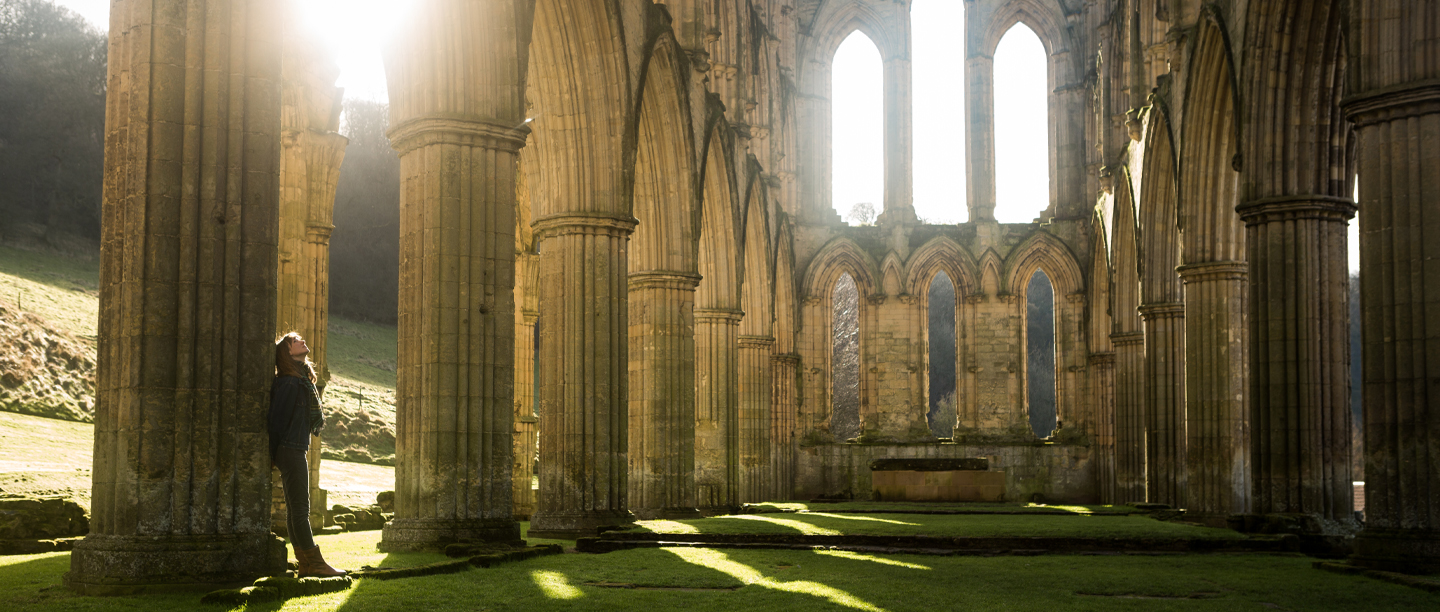 An image of the large ruins of a medieval abbey. The photo is taken from within the site's interior. The building no longer has a roof and the floor is carpeted by grass. 