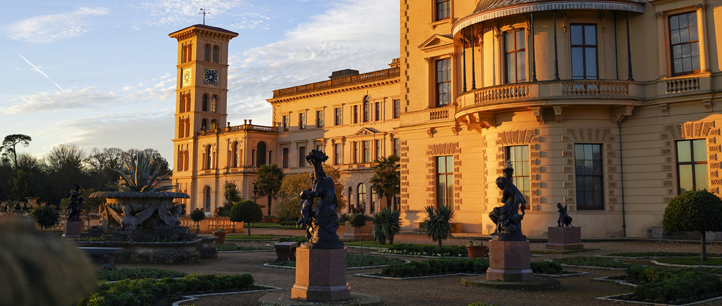 An image of the sun rising over a grand stately home. The garden in front of the house has several statues and neat flowerbeds with sculpted hedges. In  the background is a tall, rectangular, clock tower.