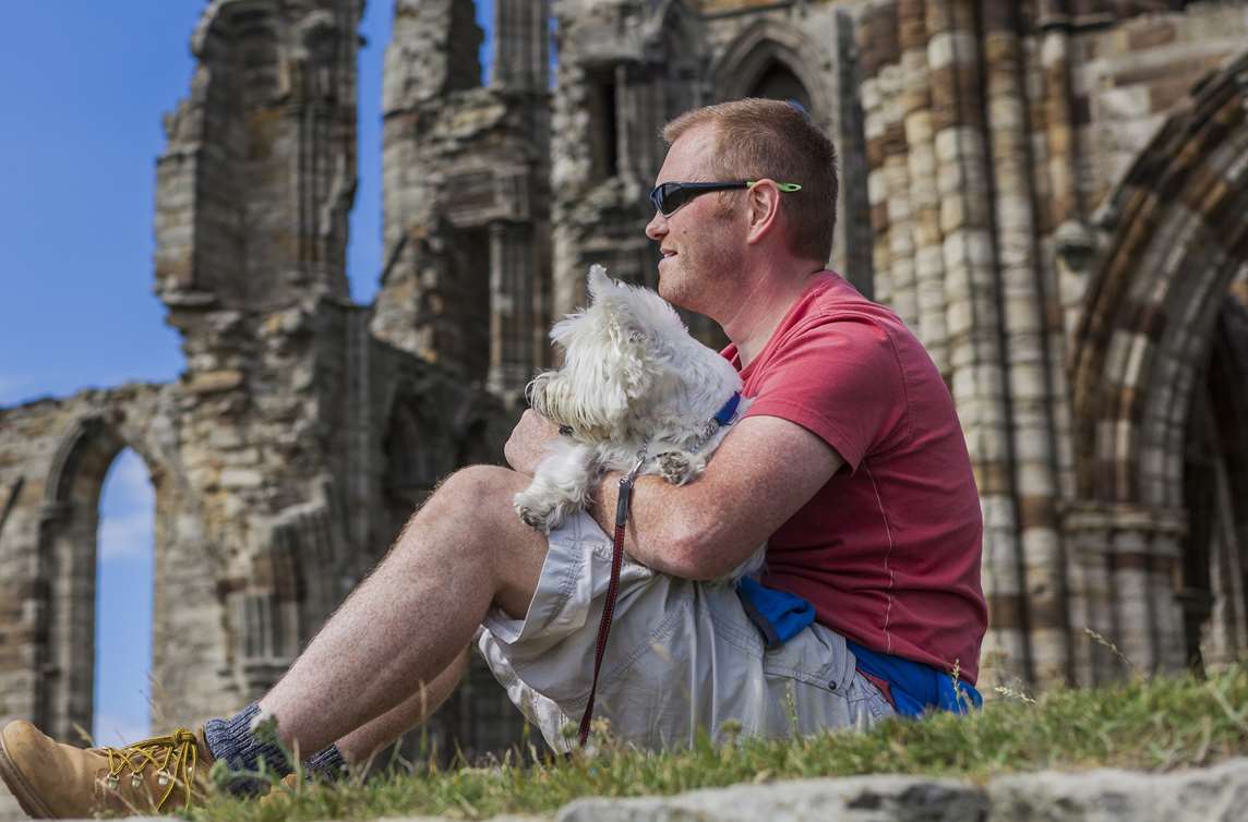 Image: A person and their dog sitting outside the ruins of Whitby Abbey