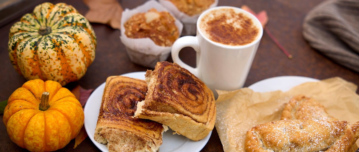 A close up of cinnamon buns, sweet pastries and muffins on a wooden table positioned next to two small pumpkins and a hot drink.