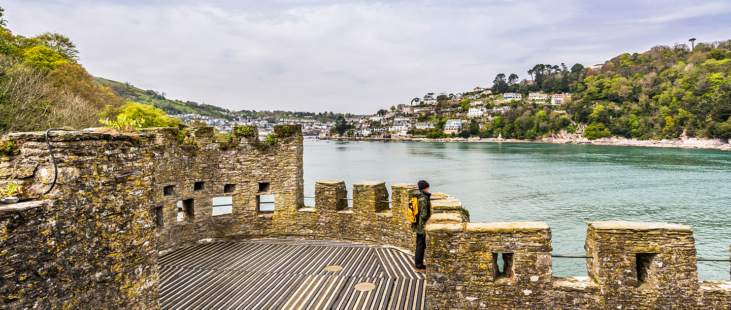 A person wearing a coat and a hat stands on a viewing platform at Dartmouth Castle.