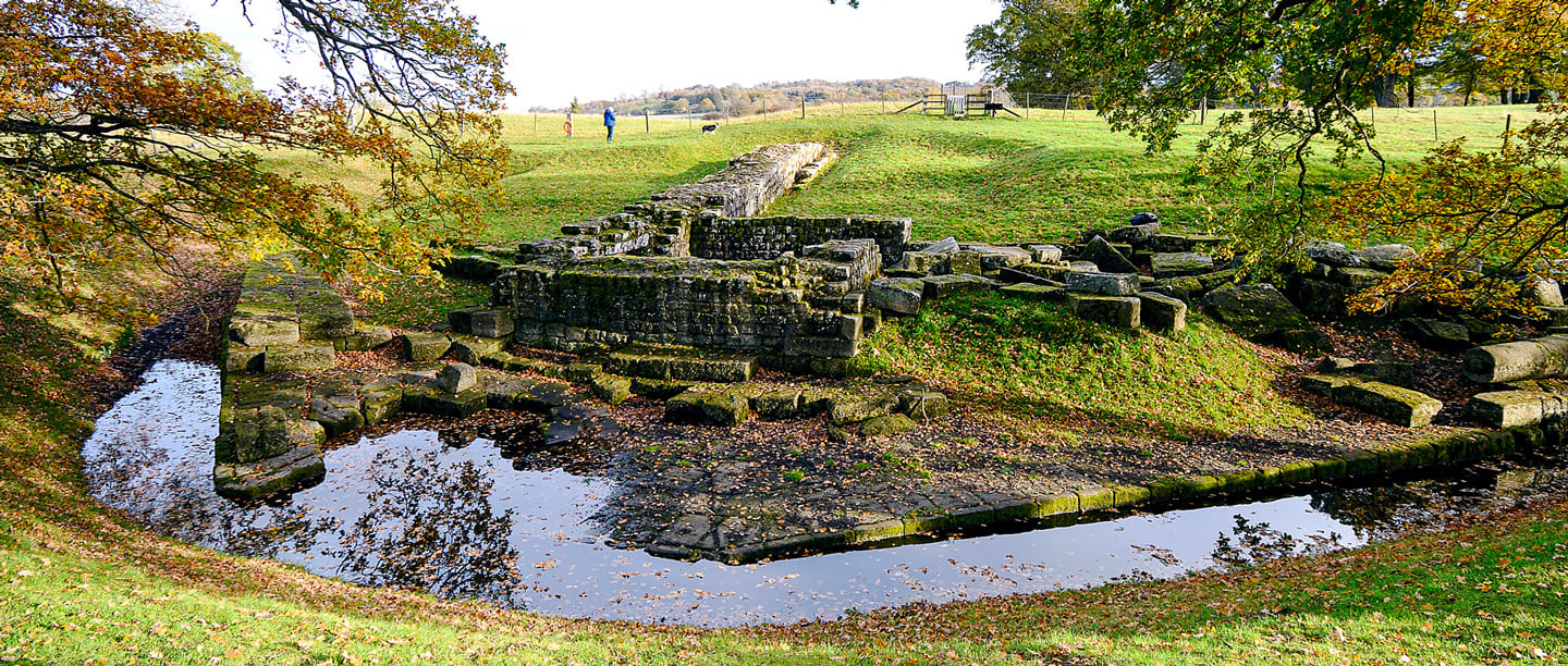The ruins of Chesters bridge abutment beside a stream and surrounded by trees and greenery.