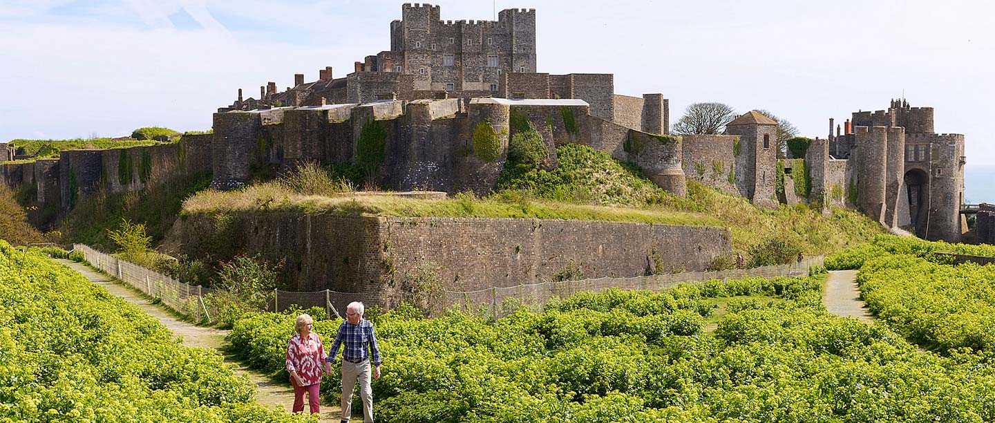 A man and a woman walk along a grass path with a huge castle behind them