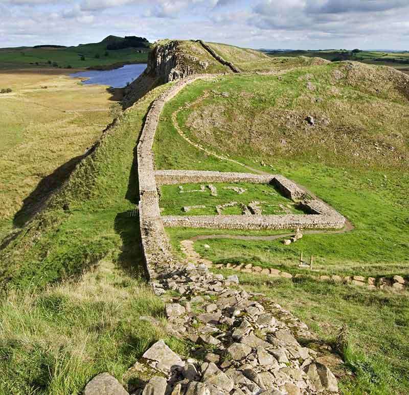 A photograph of Hadrians Wall. Half way along the remains of the outline of a square stone building can be seen.