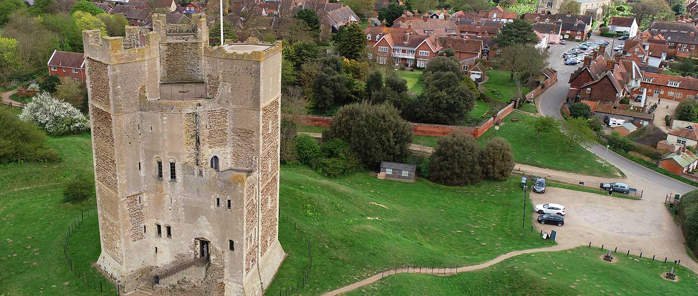 A drone shot of Orford Castle taken from almost level with the top parapets; the castle has beige stone and is on a grass hump.